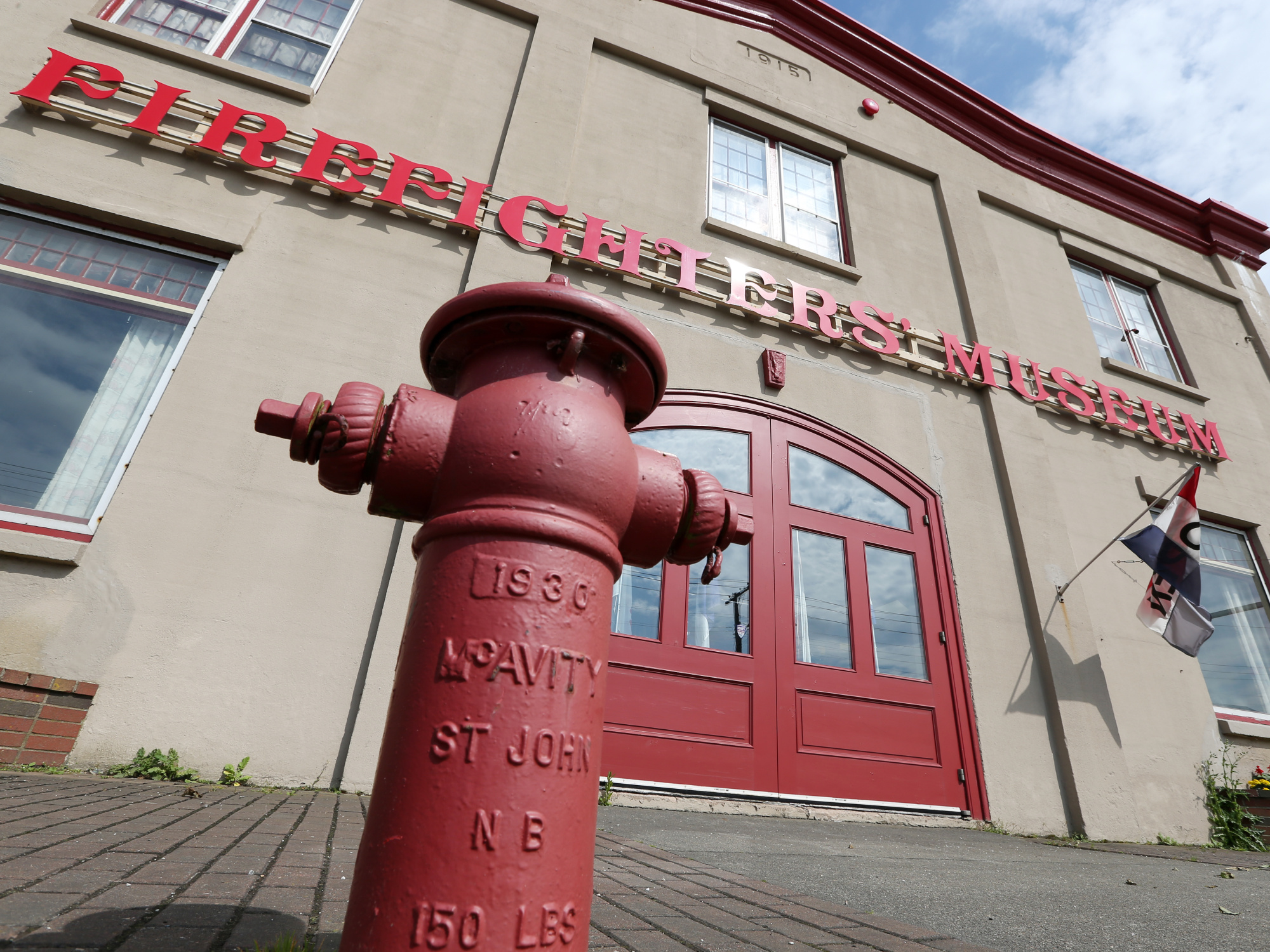 A red fire hydrant with raised lettering and numbers stands in front of a beige building with red lettering that reads "FIREFIGHTERS MUSEUM." Red doors and windows are visible on the building.