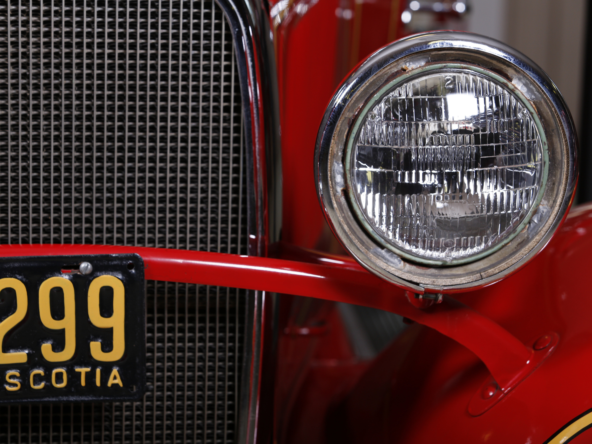 A close-up of a round chrome headlight on a red fire truck. A black license plate with yellow numbers "299" and "NOVA SCOTIA" is visible.