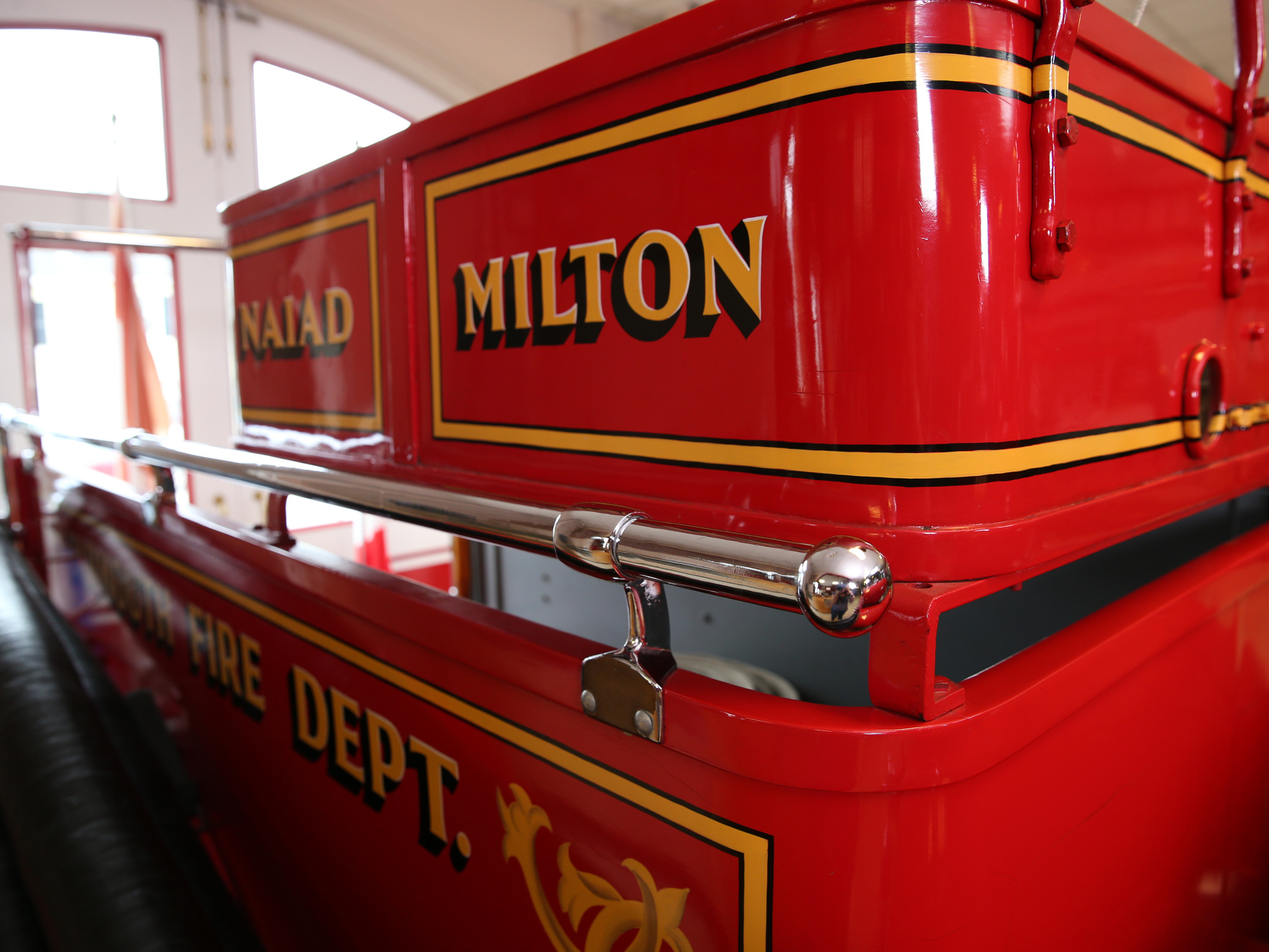 Side of a vintage firetruck. The side of the truck has "MILTON" painted in gold with black outlines, and below it, partially visible, are the words "FIRE DEPT." The truck features yellow and black pinstriping, and a polished silver railing runs along its side.